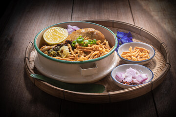 Beautiful still life of traditional Northern Thai Khao Soi chicken curry noodles served in an enamel bowl on a wooden table, with crispy noodles, shallots, lime, chili oil.