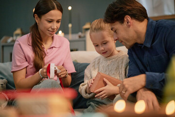 Caucasian young woman, man and child sitting together, smiling and unwrapping gift box, engaging in...