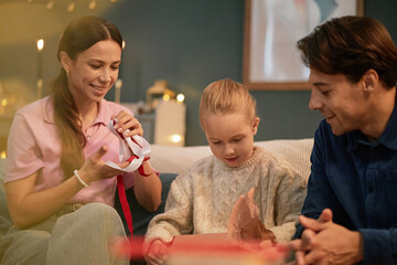 Caucasian woman and man watching young girl unwrapping gift box, all sitting together on sofa,...