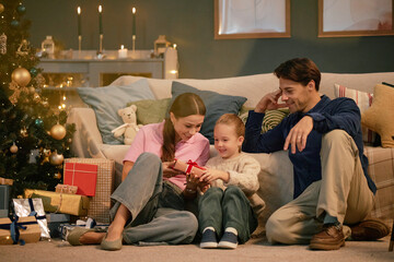 Caucasian woman and man sitting on floor with child opening gift box near decorated Christmas tree,...