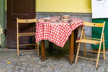 Table with red white checkered tablecloth, chained bowls and wooden chairs on cobblestone street referencing Poland’s PRL era dining practices in city