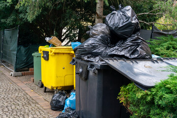 Overflowing waste bins with black trash bags, yellow recycling container, urban municipal waste area next to sidewalk surrounded by greenery