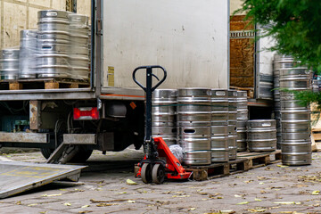 Unloading beer kegs from delivery truck, red pallet jack beside metal barrels, autumn leaves on pavement, restaurant supply area in urban environment