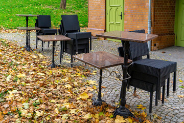 Autumn leaves covering cobblestone near outdoor café tables and rattan chairs, closed seating area in park beside brick building