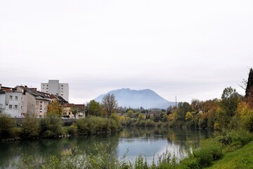 A tranquil European autumn scene with colorful fall foliage on trees along a calm river, reflecting the houses of a quiet town and a distant mountain shrouded in mist under an overcast sky.