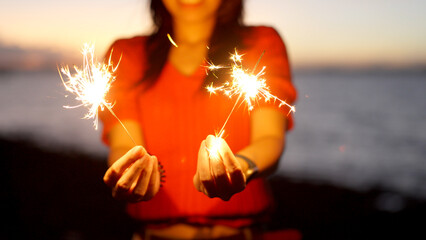 A smiling woman in a red shirt holds sparklers by the sea during sunset.