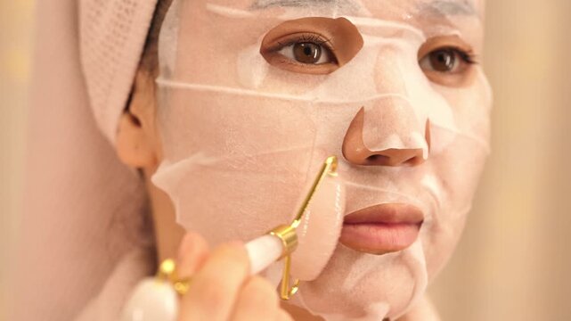 Woman in bathrobe with towel on head applying facial mask and massaging face with jade roller &mdash; skincare and beauty routine