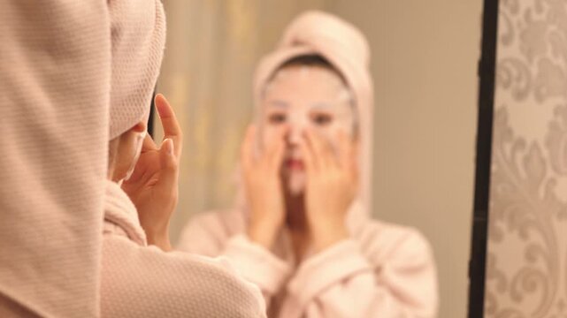 Woman in bathrobe with towel on head applying facial mask and massaging face with jade roller &mdash; skincare and beauty routine