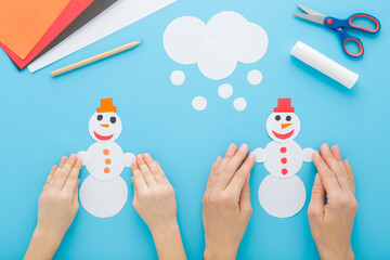 Adult mother and little child hands together making and decorating snowman shapes from white paper on blue table background. Pastel color.  Closeup. Creating winter element. Flat lay. Top down view.