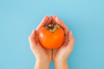 Young adult woman opened palms holding and showing ripe orange persimmon on light blue table background. Pastel color. Fresh healthy fruit. Closeup. Point of view shot. Top down view.