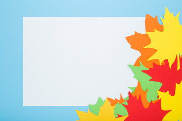 Blank white greeting card and colorful different paper leaves from application paper on light blue table background. Pastel color. Closeup. Empty place for text. Top down view.