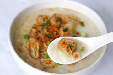 A closeup view of rice porridge in a white bowl, with a spoon scooping a portion. Generously topped with crispy fried dough (Youtiao, Yu char kway, Cakwe), chopped green onions, and fried garlic.