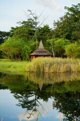 The structure is situated by a calm body of water, creating a perfect, mirrored reflection of the pavilion, surrounding trees, and the sky on the water's surface.