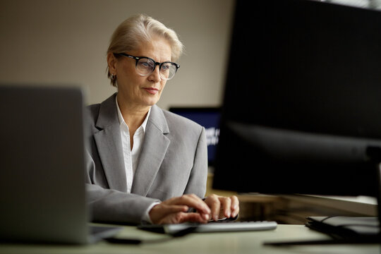 Senior Caucasian woman wearing eyeglasses working at computer in modern office, typing on keyboard and focusing on monitor, demonstrating professional business environment