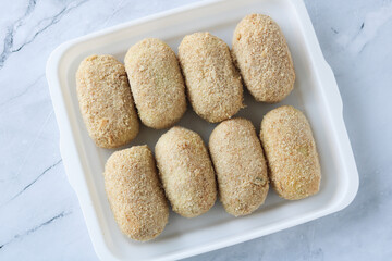 A neat row of uncooked, breaded Croquette or Kroket Ragout arranged in a white tray, ready for frying. Ideal for illustrating frozen food products or home cooking preparations.