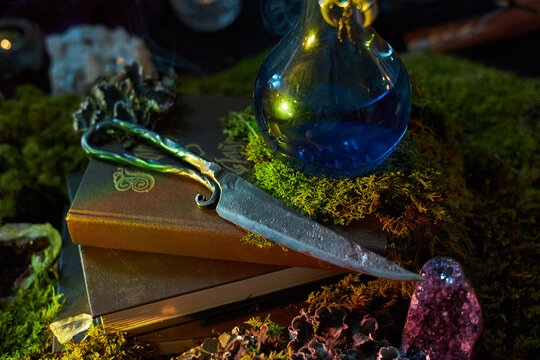 dark, enchanting scene features a silver dagger resting on an ancient book. blue potion glimmers nearby, surrounded by moss and mystic crystals, creating a magical atmosphere