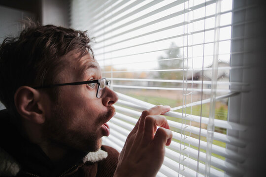 Surprised man in glasses peeking through window blinds. Spying on the neighbors.