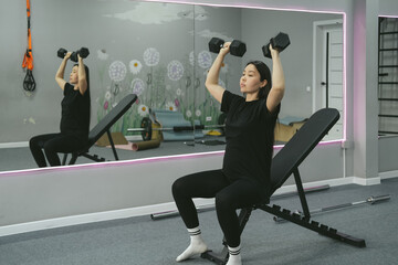 Strong beautiful fitness asian woman training, doing exercise with dumbbells at gym. Female sport athlete coach making workout bodybuilding on bench heavy fit equipment.