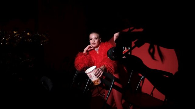 Portrait of a beautiful young woman in a red fur coat against a red background, posing for a cinema-style photo. The woman smiles beautifully, sitting on chairs in the light of a projector during a