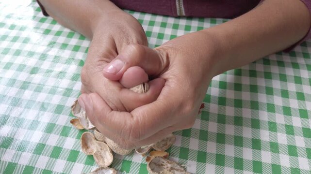 Close-up shot of person cracking peanut shells and peeling nuts on green checkered tablecloth.