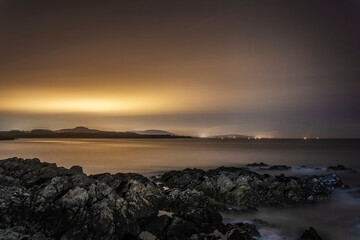 Atmospheric nocturnal seascape showing ethereal water, rocky shore, and amber light pollution in the sky