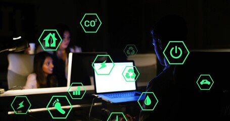 Typing man wearing t-shirt on laptop in office at night, with floating green icons, copy space
