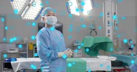 Female surgeon standing in operating room wearing blue gown, mask, cap with floating hexagon icons