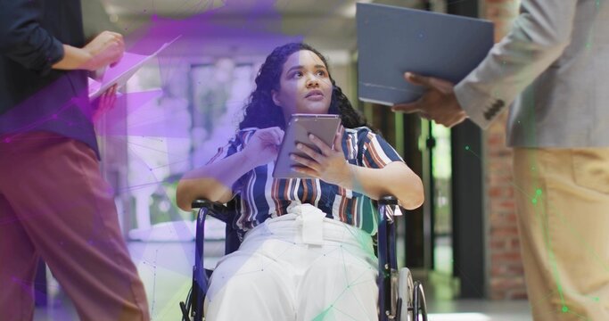 Gesturing wheelchair user showing tablet to colleagues with clipboard and laptop in office corridor - Powered by Adobe