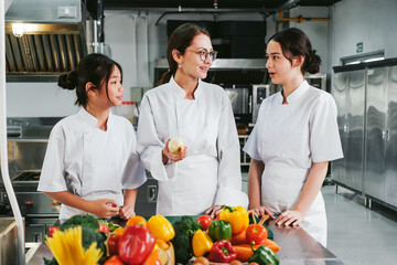 Three chefs in white uniforms discuss prep in a professional commercial kitchen, surrounded by colorful fresh vegetables on a stainless counter, conveying teamwork, training, and culinary planning.