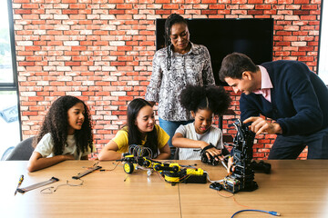 Students build robots with guidance from instructors at a classroom table, using wires, tools, and...