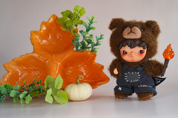 Adorable doll dressed in a fluffy bear costume holds a small torch with autumn-themed decorations nearby. Vibrant orange flowers and pumpkins accent the cozy seasonal setup.