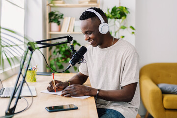 Young African journalist taking notes while recording live podcast in home studio. Focused content...