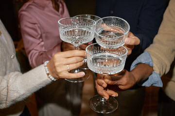 Diverse group of young adult men and women clinking crystal glasses in celebratory toast, hands and arms visible, multiethnic gathering, close up view