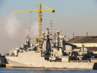 Navio de guerra ou fragata militar cinza ancorado em porto industrial, com um guindaste amarelo de torre alta ao fundo. A cena sugere manuten&ccedil;&atilde;o naval ou atividade de base militar.