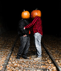 Couple wearing pumpkin heads share a playful moment on a leaf-covered train track in autumn twilight