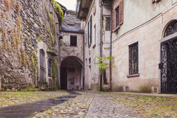 Narrow stone alley on San Giulio Island in Lake Orta, ancient walls covered with moss and iron gates leading to arched passage beneath soft diffused light