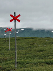 Red winter trail markers standing out against lush green mountain terrain and low clouds in the Swedish highlands &mdash; a symbol of guidance and solitude in the Scandinavian wilderness.