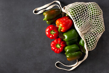 Colorful red and green bell peppers in a reusable mesh bag on a dark stone background. Eco-friendly shopping, zero waste, and organic vegetable concept for healthy sustainable lifestyle.