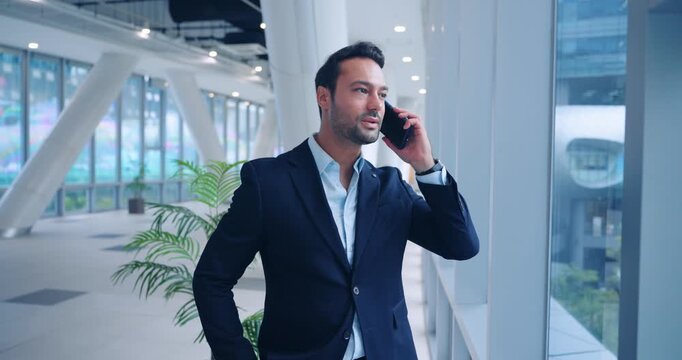 Professional male executive standing by large window talking on smartphone in bright modern interior of corporate office. - Powered by Adobe