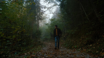 Obraz premium Man walking through a foggy forest pathway surrounded by trees and autumn leaves on a quiet day