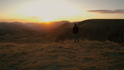 Man stands alone on a hilltop while watching the sunset in a mountainous landscape with expansive views during golden hour