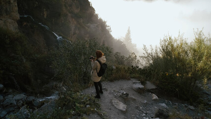 Obraz premium Woman exploring a scenic trail near a waterfall while taking photos in the early morning light at a mountain location