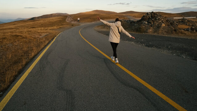 Woman enjoying a joyful moment as she balances on the yellow line of a quiet mountain road during sunset in an open landscape