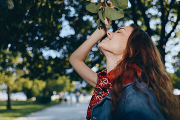 smiling woman enjoying nature with green leaves and sunny outdoor park background wearing red scarf and denim jacket in warm weather