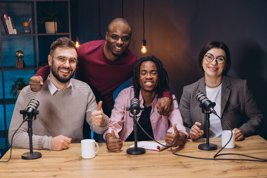 Four podcasters sitting at a table with microphones, smiling and giving thumbs up during a live recording