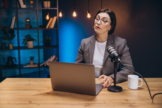 Middle aged woman recording her podcast using a laptop and a professional microphone in a home studio with blue light and bookshelves