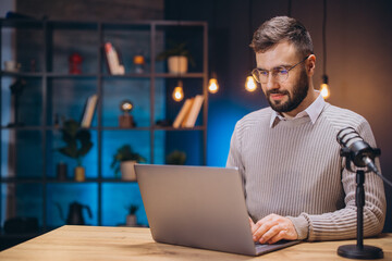 Podcaster working at home, speaking into professional microphone while using laptop for recording digital audio content