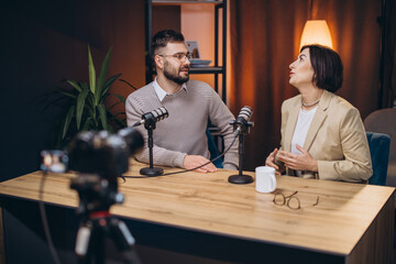 Man and woman sitting at a table with microphones and a camera, recording a podcast episode