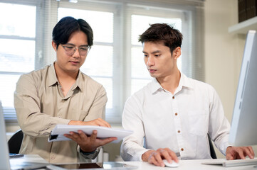 Asian businessman looking at document as sitting aside coworker at computer table in company office.
