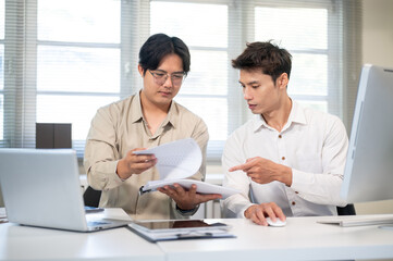 Asian businessman and coworker looking at document while sitting at computer table in company office
